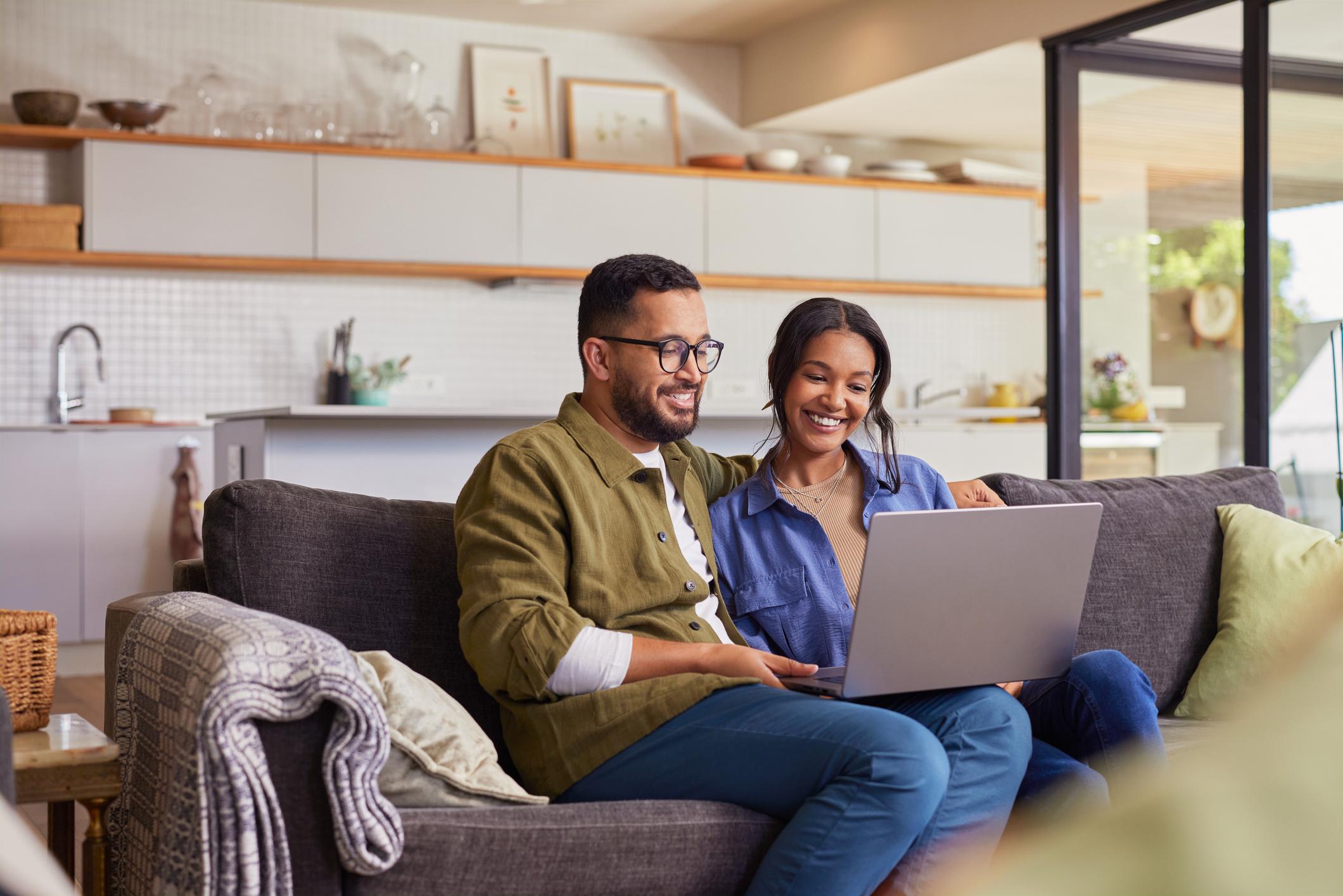Young couple watching movie on laptop while relaxing on a weekend at home. Smiling indian man and beautiful woman sitting on sofa while using laptop. Happy mid adult caual man and beautiful hispanic woman sitting on couch while surfing the net.