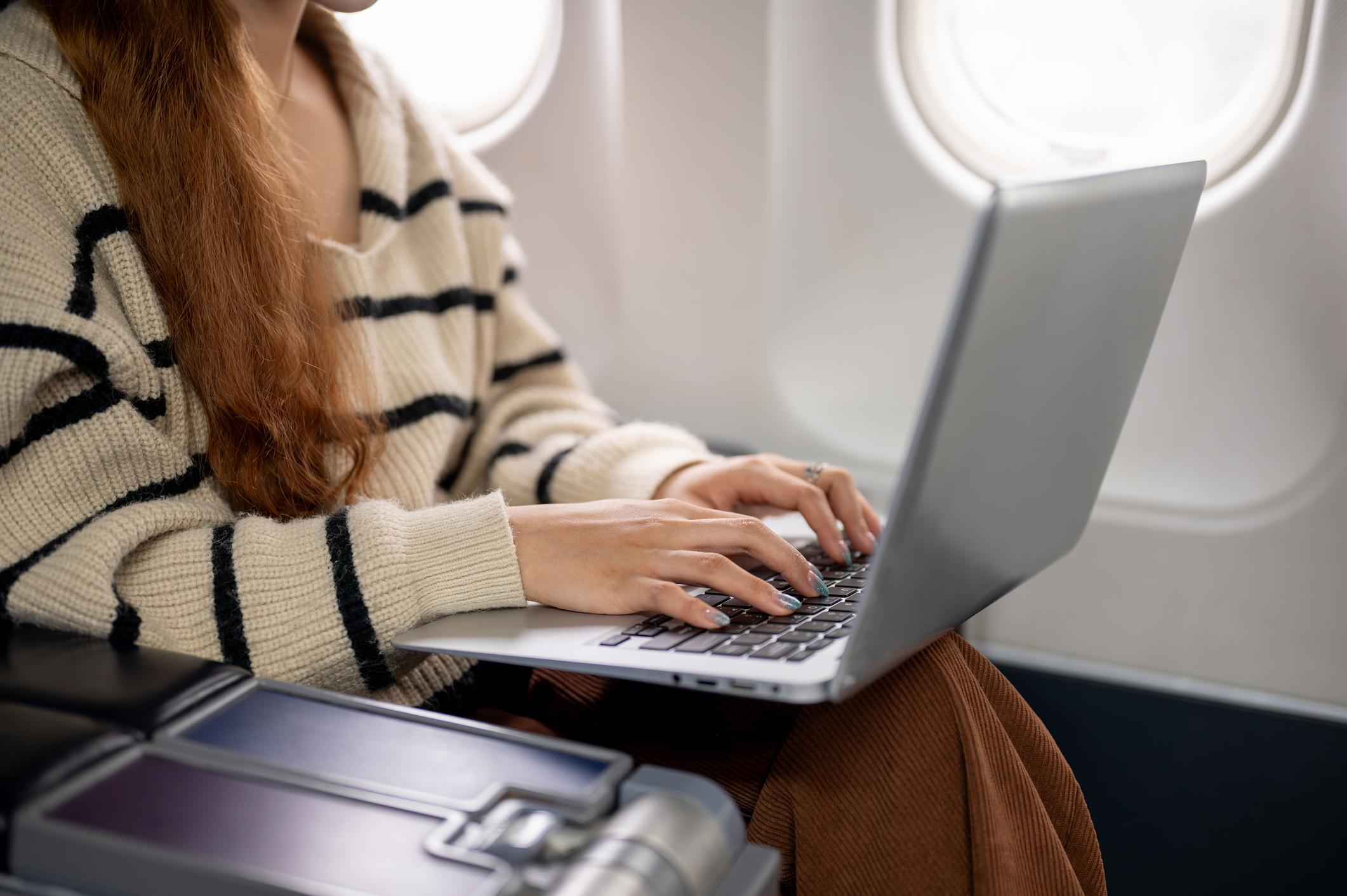 A close-up side view of a woman in cozy clothes sitting at a window seat, working on her laptop and typing on the keyboard during the flight, traveling to somewhere.