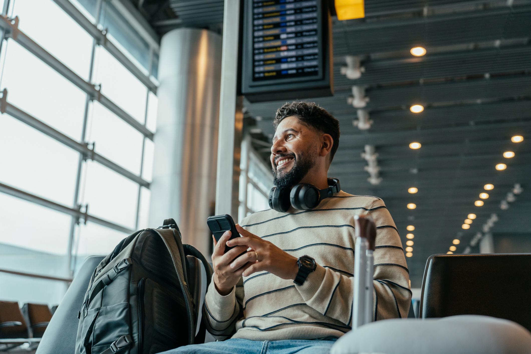 A male tourist holding his smartphone and smiling while waiting to board at the airport.