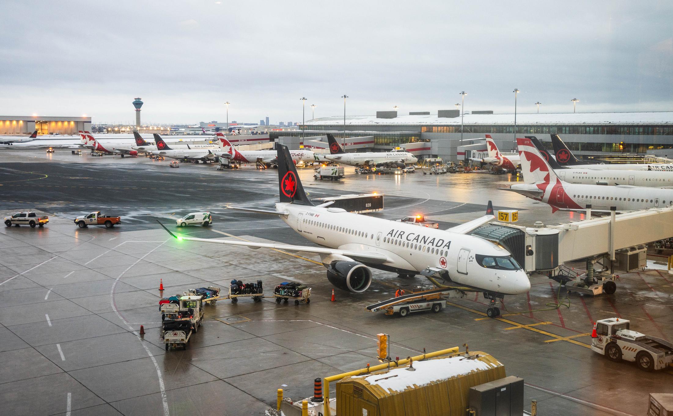 An air canada airbus a220 jet stands at a boarding gate during a rainy winter day at toronto pearson international airport (yyz). the wet tarmac reflects the airport lights as ground crew vehicles and other aircraft, including air canada rouge and porter airlines, are seen in the background. this editorial image captures the daily operations and winter aviation atmosphere at canada's busiest transportation hub in february 2026.