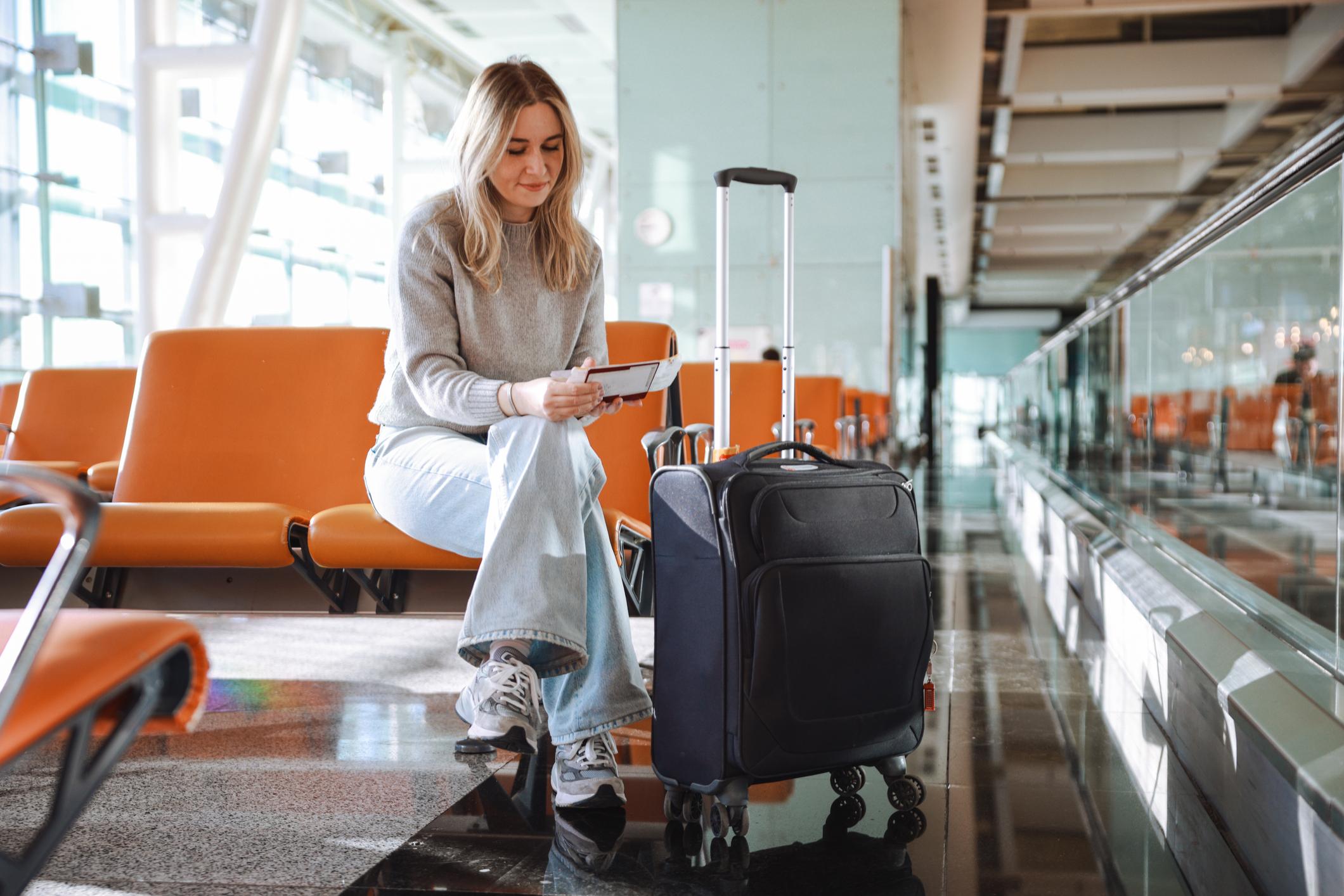 A woman waits at the airport, looking aside excited and holding her boarding pass and passport, with a bag close by.
