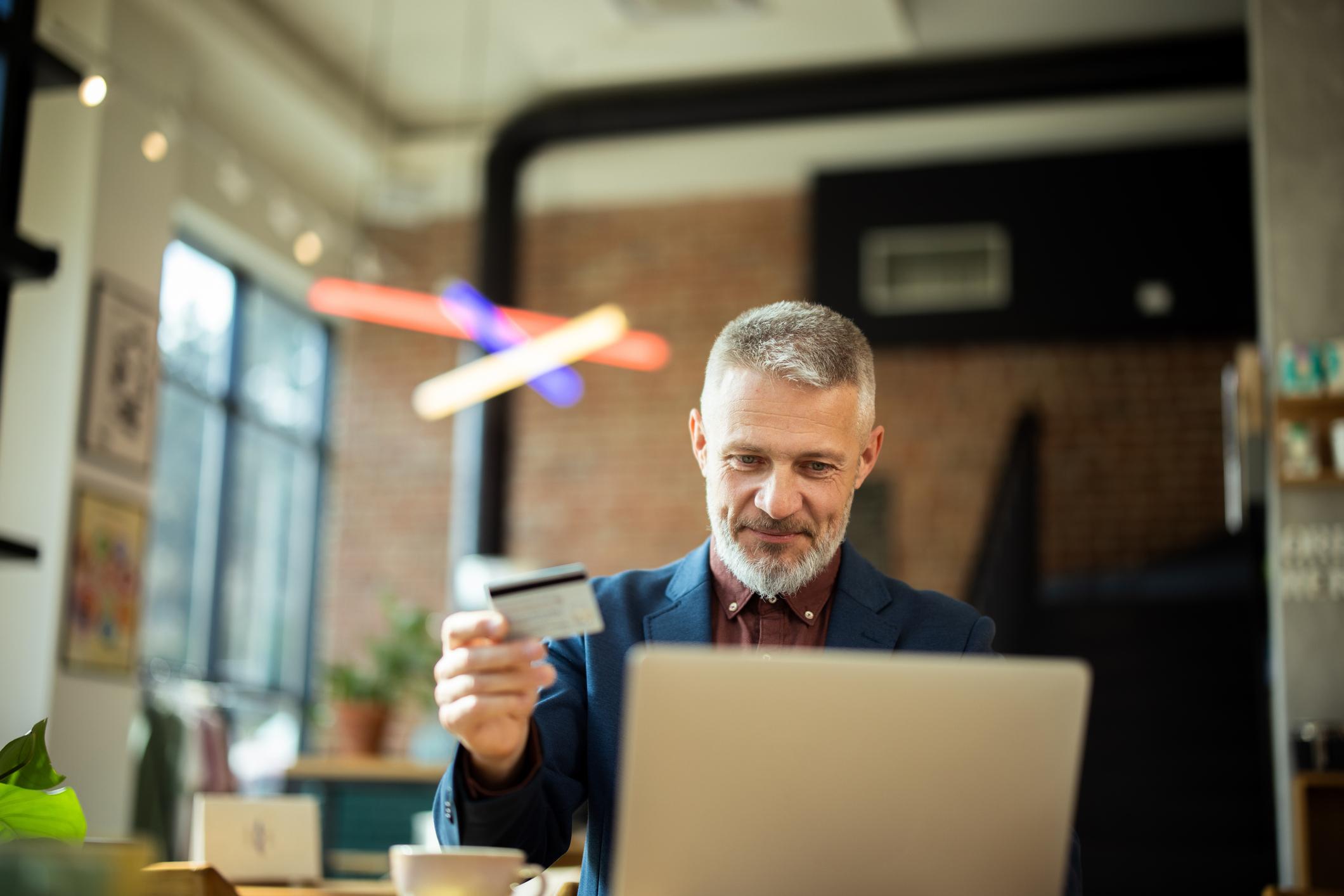 Focused mature businessman is holding a credit card and making an online payment on a laptop in a cafe