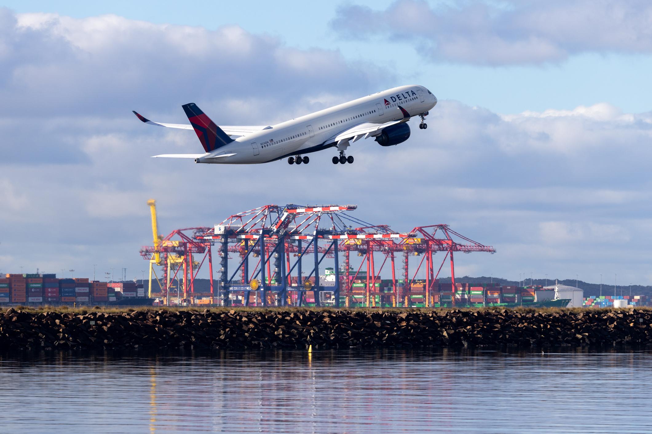 A Delta Air Lines Airbus A350-941 plane, registration N514DN, taking off from Sydney Kingsford Smith Airport as flight DL40 to Los Angeles. In the background are cranes, shipping containers, fuel storage tanks and two container ships at the Port Botany Container Terminal. This image was taken from near Tower Beach, Botany Bay on a cold, sunny and partly cloudy morning on 18 July 2026.
