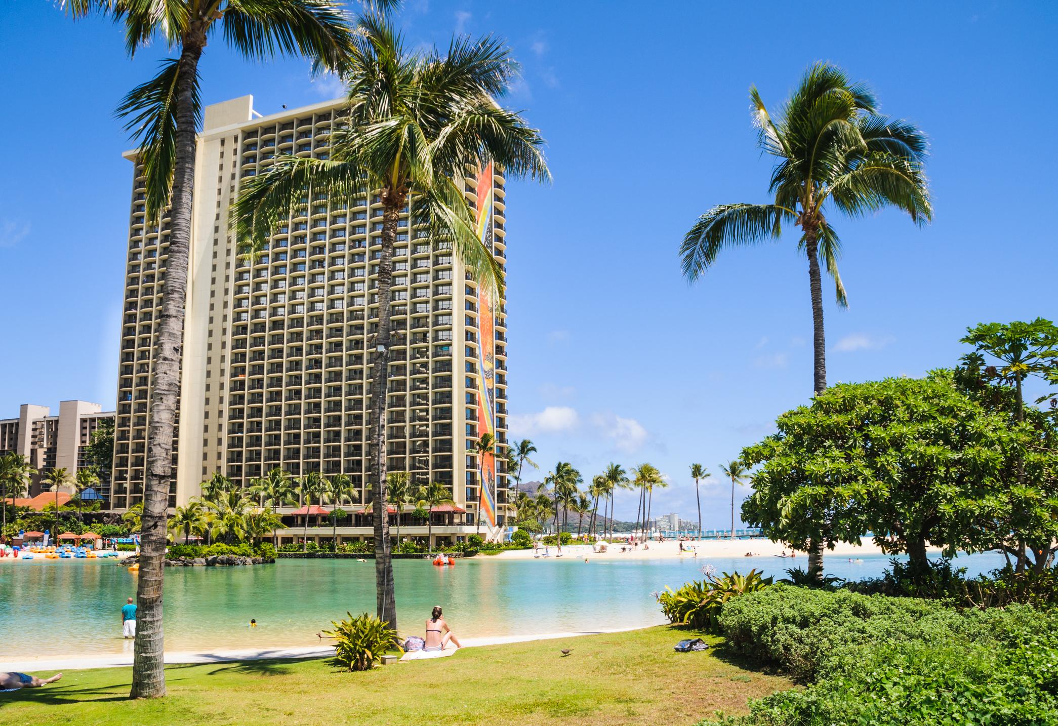 A young woman sits on a blanket on the grass fringe of the Duke Kahanamoku Lagoon watching others swim and paddle its shallow waters.