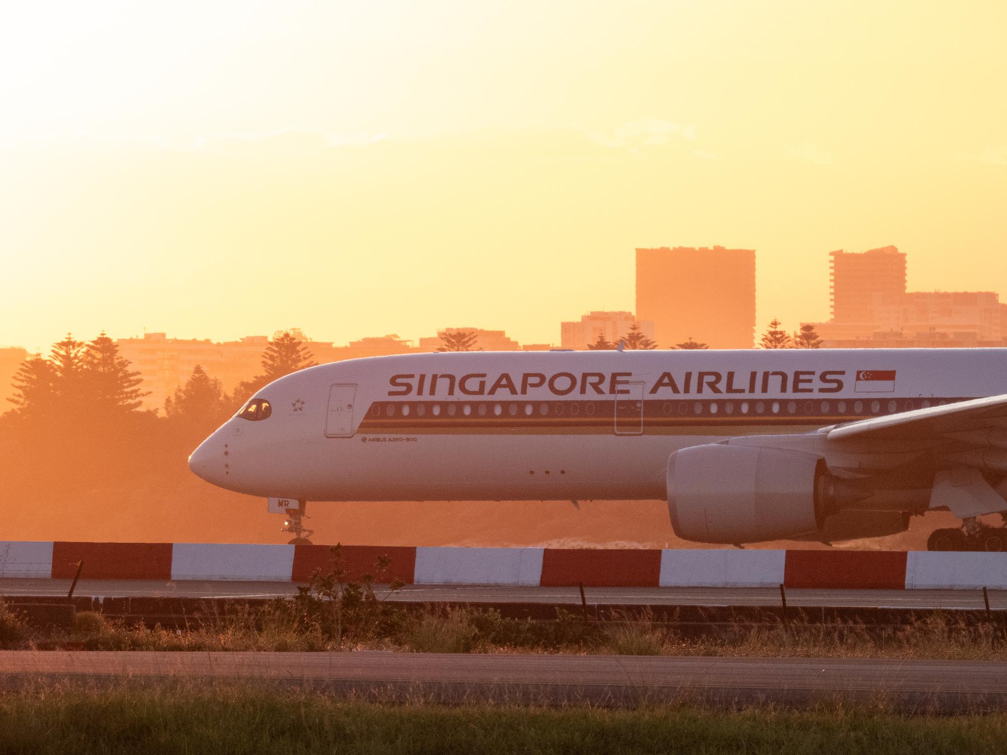 A Singapore Airlines Airbus A350-941 plane, registration 9V-SMR, taxiing at Sydney Kingsford Smith Airport in preparation for departure as flight SQ242 to Singapore. The red and white striped barrier marks the main runway. This image was taken from Shep's Mound, Ross Smith Avenue, Mascot on a hot and sunny evening approaching sunset on 13 December 2024.