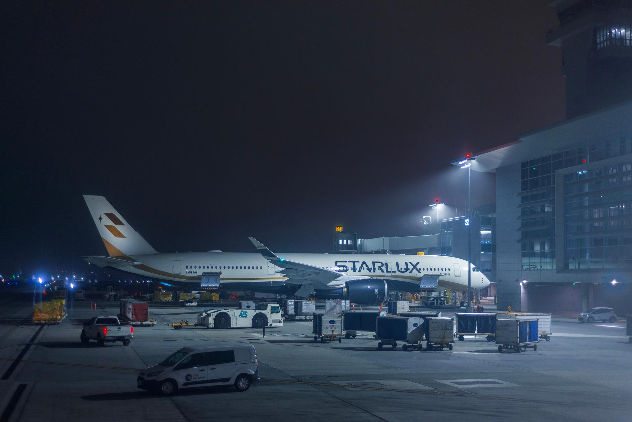 STARLUX Airlines aircraft parked beside an airport terminal during nighttime ground operations surrounded by baggage handling vehicles. Light mist and artificial illumination create an atmospheric scene highlighting aviation logistics, passenger transportation, airline operations, airport infrastructure and global air travel at Dallas Fort Worth International Airport.