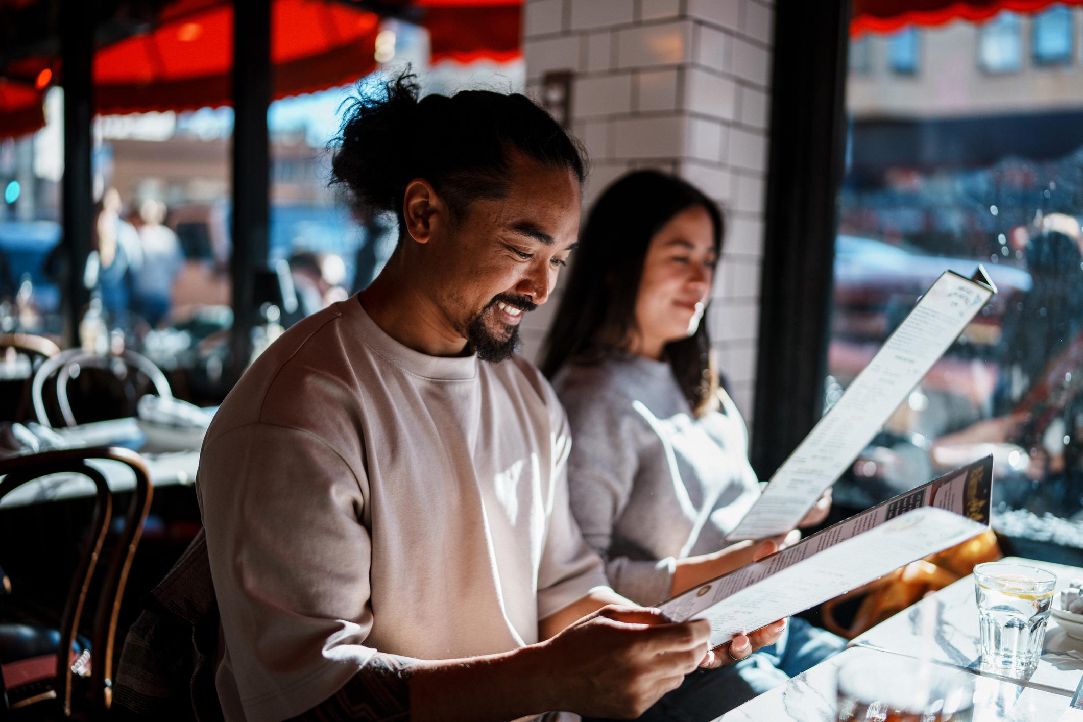 A Filipino couple in their 30s who are enjoying a fun and relaxing weekend staycation exploring San Francisco, California, smile while reviewing the menu at a restaurant in the Little Italy neighborhood of the city.